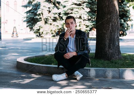 A Young Guy Smokes Near A Tree In The City. A Black Jacket, Jeans, White Sneakers, A White T-shirt A