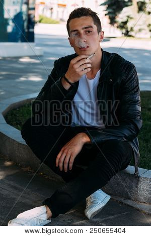 A Young Guy Smokes Near A Tree In The City. A Black Jacket, Jeans, White Sneakers, A White T-shirt A