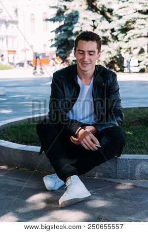 A Young Guy Smokes Near A Tree In The City. A Black Jacket, Jeans, White Sneakers, A White T-shirt A