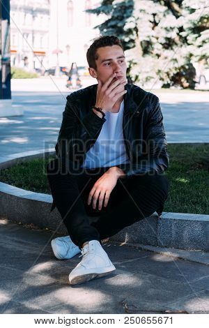 A Young Guy Smokes Near A Tree In The City. A Black Jacket, Jeans, White Sneakers, A White T-shirt A