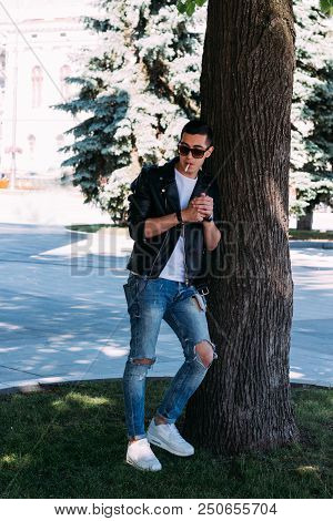 A Young Guy Smokes Near A Tree In The City. A Black Jacket, Jeans, White Sneakers, A White T-shirt A