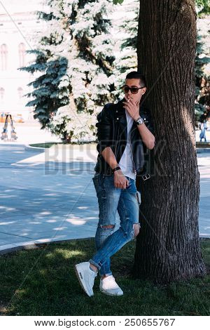 A Young Guy Smokes Near A Tree In The City. A Black Jacket, Jeans, White Sneakers, A White T-shirt A