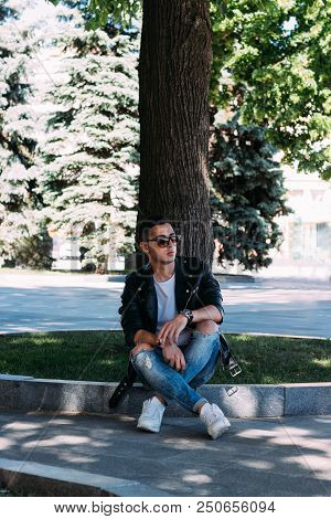 A Young Guy Smokes Near A Tree In The City. A Black Jacket, Jeans, White Sneakers, A White T-shirt A