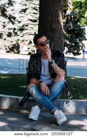 A Young Guy Smokes Near A Tree In The City. A Black Jacket, Jeans, White Sneakers, A White T-shirt A