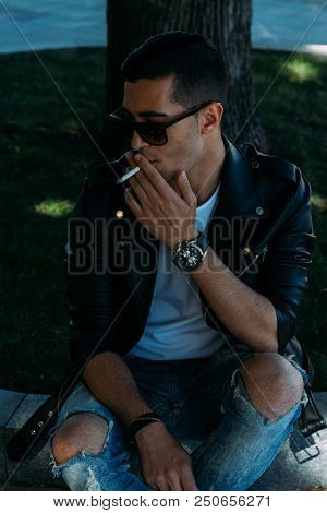 A Young Guy Smokes Near A Tree In The City. A Black Jacket, Jeans, White Sneakers, A White T-shirt A