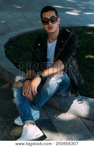 A Young Guy Smokes Near A Tree In The City. A Black Jacket, Jeans, White Sneakers, A White T-shirt A