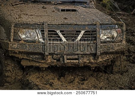 Part Of Offroad Car Stuck In Deep Mud On Dark Background. Bonnet, Lights And Bumper Covered With Dir