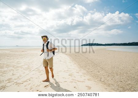 Free Travel Male Hiking On A Tropical Beach Barefoot With A Backpack In A White Shirt, Hat, Shorts A