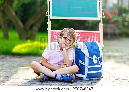 Frustrated Sad Little Kid Boy With Glasses Sitting By Desk And Backpack Or Satchel. Schoolkid With T