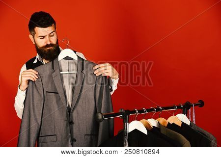 Man Looks For Clothes At Fashion Shop. Bearded Hipster Man Stands Near Rack With Clothes Against Red