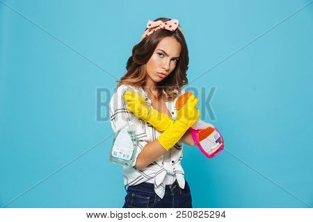 Portrait closeup of concenteated serious woman 20s in yellow rubber gloves for hands protection holding two detergent sprayers during cleaning isolated over blue background