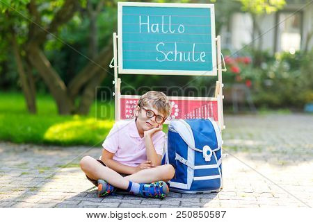 Frustrated Sad Little Kid Boy With Glasses Sitting By Desk And Backpack Or Satchel. Schoolkid With T
