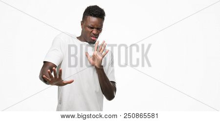Young african american man wearing white t-shirt disgusted expression, displeased and fearful doing disgust face because aversion reaction. With hands raised. Annoying concept.