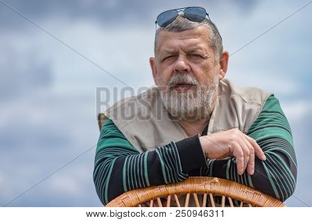 Outdoor Portrait Of Senior Man In Black Sunglasses Leant On A Wicker Chair Against Cloudy Sky