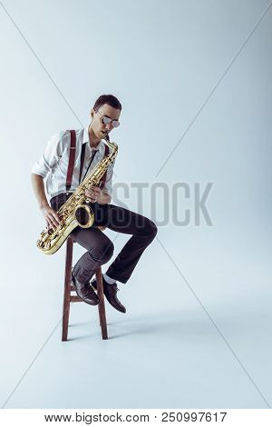Handsome Young Jazzman Sitting On Stool And Playing Saxophone On Grey