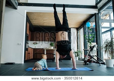 Father Exercising On A Yoga Mat, Staning On His Hands Next To His Infant Baby