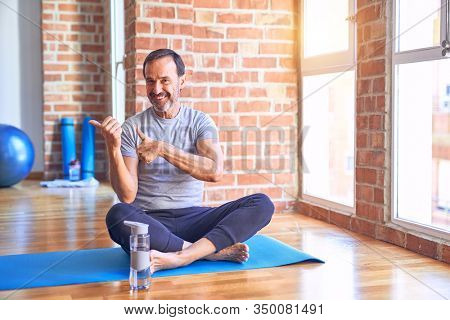 Middle age handsome sportman sitting on mat doing stretching yoga exercise at gym Pointing to the back behind with hand and thumbs up, smiling confident
