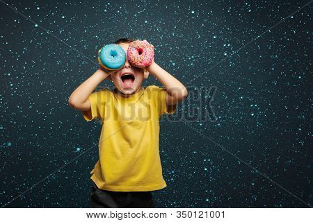 Happy cute boy is having fun played with donuts on black background wall. Bright photo of a boy. Colored donuts