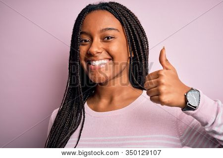 Close up of young african american woman wearing pink sweater over isolated background doing happy thumbs up gesture with hand. Approving expression looking at the camera showing success.