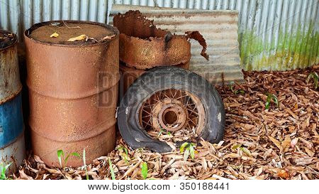 Old Rusted Forty Four Gallon Drums And A Car Spoke Tyre Lying Outdoor Amongst Leaves, On Display As 