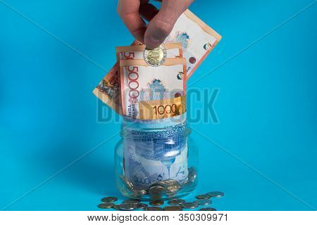 The Hand Puts A Tenge Coin In A Glass Jar With Money. Kazakhstan. Kz. Kzt. Banknote, Paper Currency,