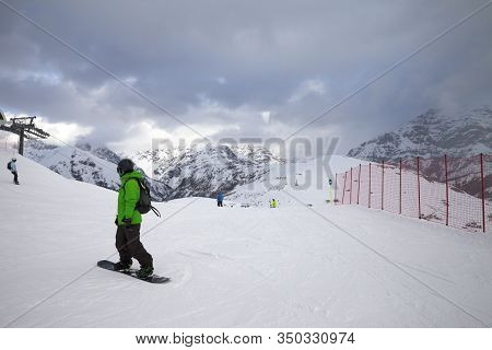 Snowy Ski Slope With Snowboarders And Skiers In High Mountains And Cloudy Sunlit Sky At Winter Gray 