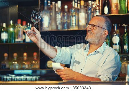 Elderly Bartender Cleans The Glass. A Handsome Bartender Polishes A Glass Of Wine Glasses. The Conce