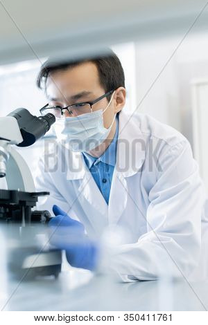Vertical portrait of young Asian pharmacologist wearing protective mask examining specimen chemical reaction using microscope in modern laboratory
