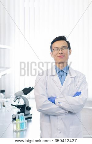 Confident Asian pharmacologist wearing white lab coat and eyeglasses standing with arms crossed looking at camera smiling in modern laboratory
