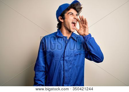 Young mechanic man wearing blue cap and uniform standing over isolated white background shouting and screaming loud to side with hand on mouth. Communication concept.