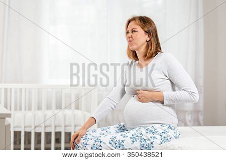 A Young Pregnant Woman Is Sitting On The Bed And Writhing With Pain In Her Belly. Childrens Room In 
