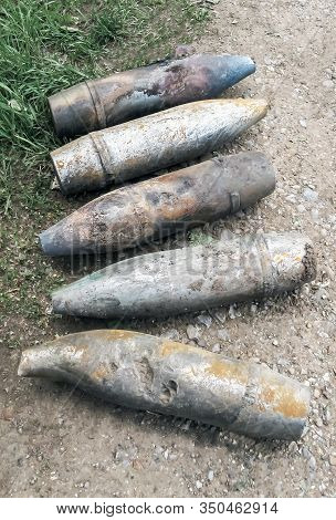 Five Unexploded Artillery Shells Lie On The Side Of The Asphalt Road, Dandelions Bloom And The Grass