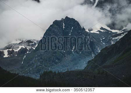 Bleak Dramatic Alpine Landscape With Big Cliff In Low Cloud Near Giant Snowy Mountain Wall In Dark E