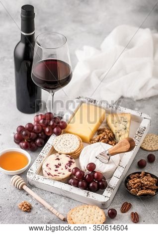 Glass And Bottle Of Red Wine With Selection Of Various Cheese In Wooden Box And Grapes On Light Back