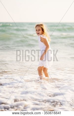 A Blonde Girl In A White Dress Standing In The Sea Knee-deep. Waves At Sea