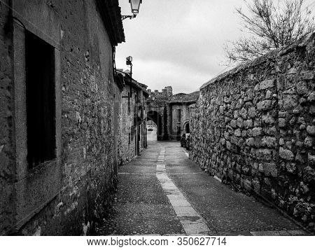 Black And White Photo Of A Narrow Street Surrounded By Stone Walls With A Stone Tunnel In The Backgr