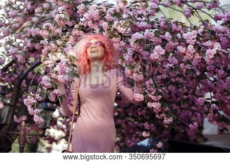 Positive Young Woman With Curly Pink Hair And Sunglasses Near The Blossoming Spring Tree.