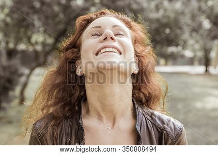 Happy Pretty Young Lady Throwing Head Back In City Park. Woman Wearing Leather Jacket And Keeping Ey