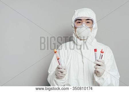 Asian Epidemiologist In Hazmat Suit And Respirator Mask Holding Test Tubes With Blood Samples On Gre