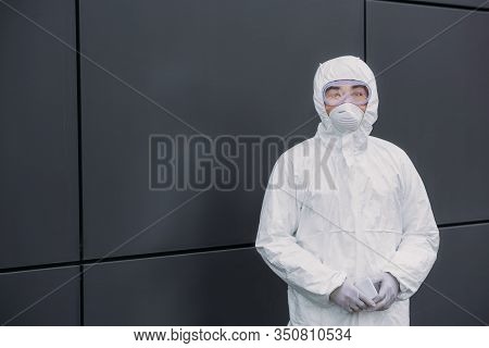 Asian Epidemiologist In Hazmat Suit And Respirator Mask Looking At Camera While Standing Near Wall