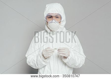 Asian Epidemiologist Looking At Camera While Holding Thermometer Showing High Temperature On Grey Ba