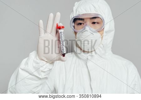 Asian Epidemiologist In Hazmat Suit And Respirator Mask Showing Test Tube With Blood Sample On Grey 