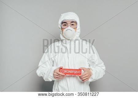 Asian Epidemiologist In Hazmat Suit And Respirator Mask Holding Warning Card With Coronavirus Inscri
