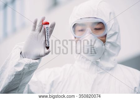 Asian Epidemiologist In Hazmat Suit And Respirator Mask Looking At Test Tube With Blood Sample While