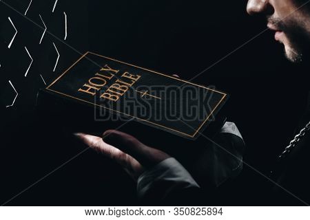 Cropped View Of Catholic Priest Holding Holy Bible Near Confessional Grille In Dark With Rays Of Lig