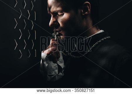 Young Tense Catholic Priest Kissing Cross On His Necklace In Dark Near Confessional Grille With Rays