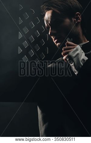 Young Serious Catholic Priest Touching Cross On His Necklace In Dark Near Confessional Grille