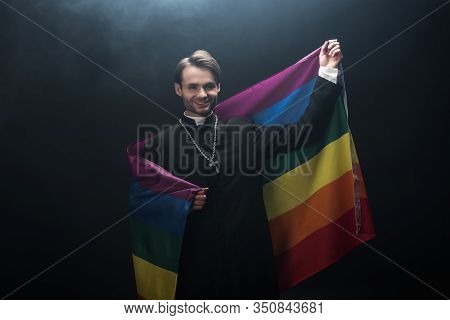 Smiling Catholic Priest Holding Lgbt Flag While Looking At Camera On Black Background With Smoke