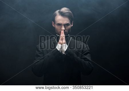 Serious Catholic Priest Looking At Camera While Praying On Black Background With Smoke