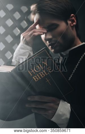 Young Serious Catholic Priest Touching Face While Reading Bible Near Confessional Grille In Dark Wit